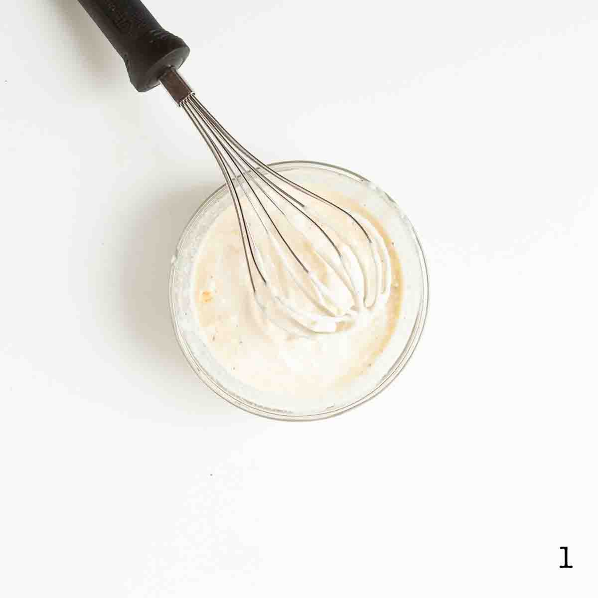 A whisk resting in a bowl of Caesar dressing.