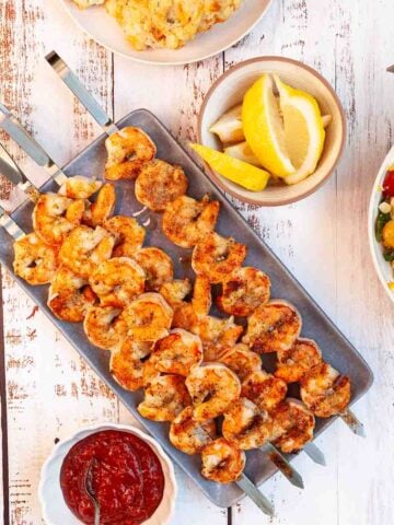 Four grilled shrimp skewers on a platter with lemon wedges and cocktail sauce on the side.