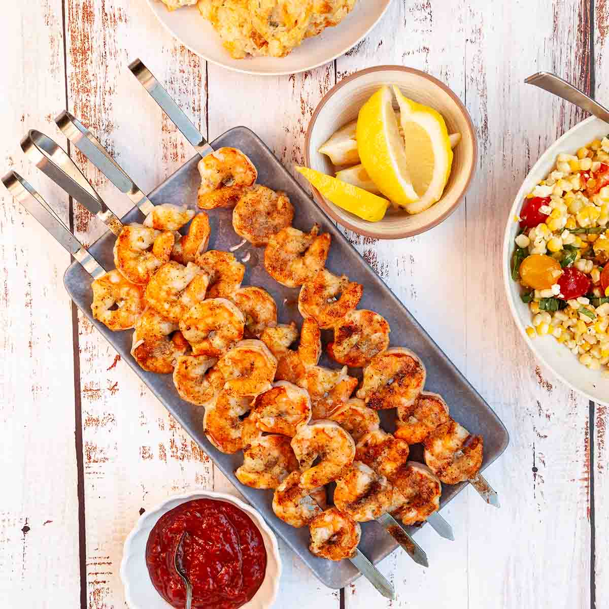 Four grilled shrimp skewers on a platter with lemon wedges and cocktail sauce on the side.