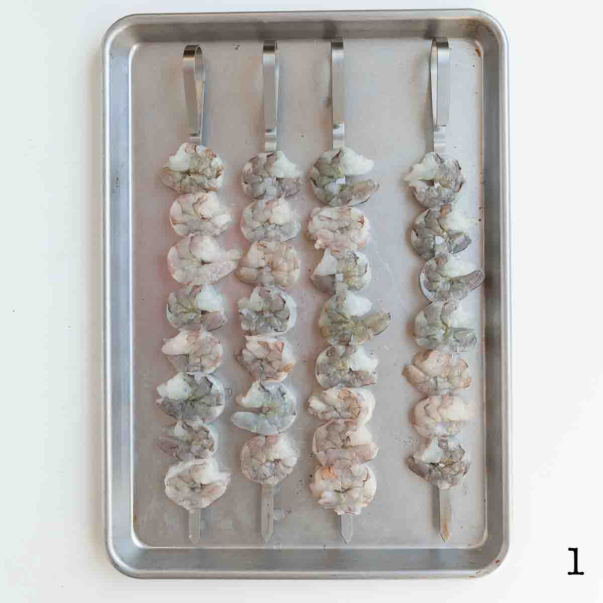 Four skewers of raw shrimp on a metal baking sheet.