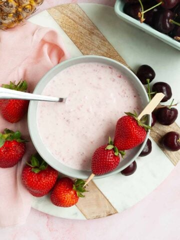 A bowl of strawberry fruit dip with strawberries and cherries scattered around it.