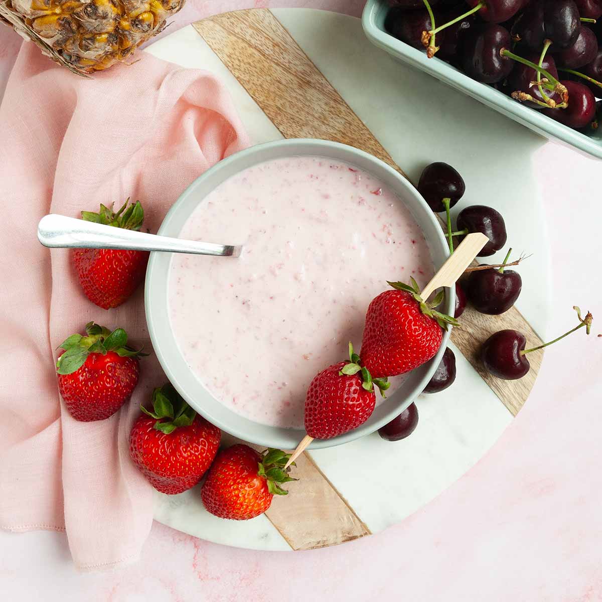 A bowl of strawberry fruit dip with strawberries and cherries scattered around it.