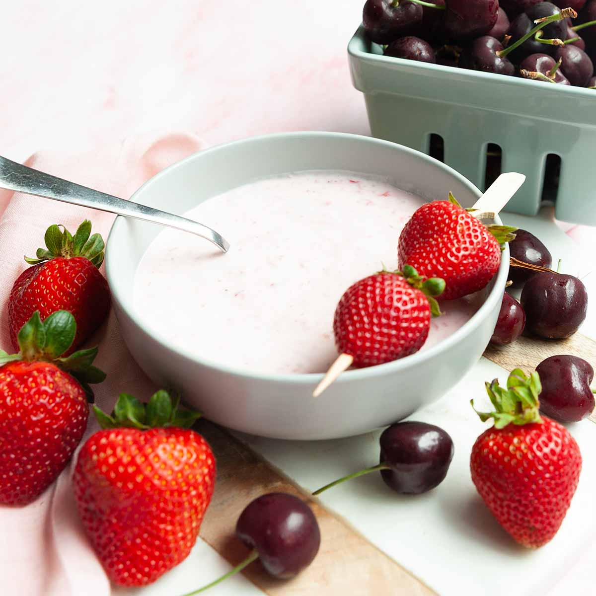 A bowl of strawberry fruit dip with strawberries and cherries scattered around it.