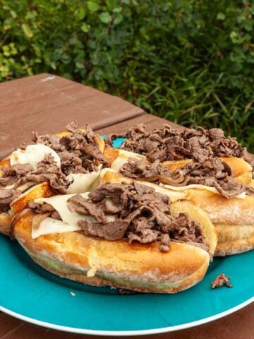 Four Blackstone cheesesteaks on a large plate on a picnic table.