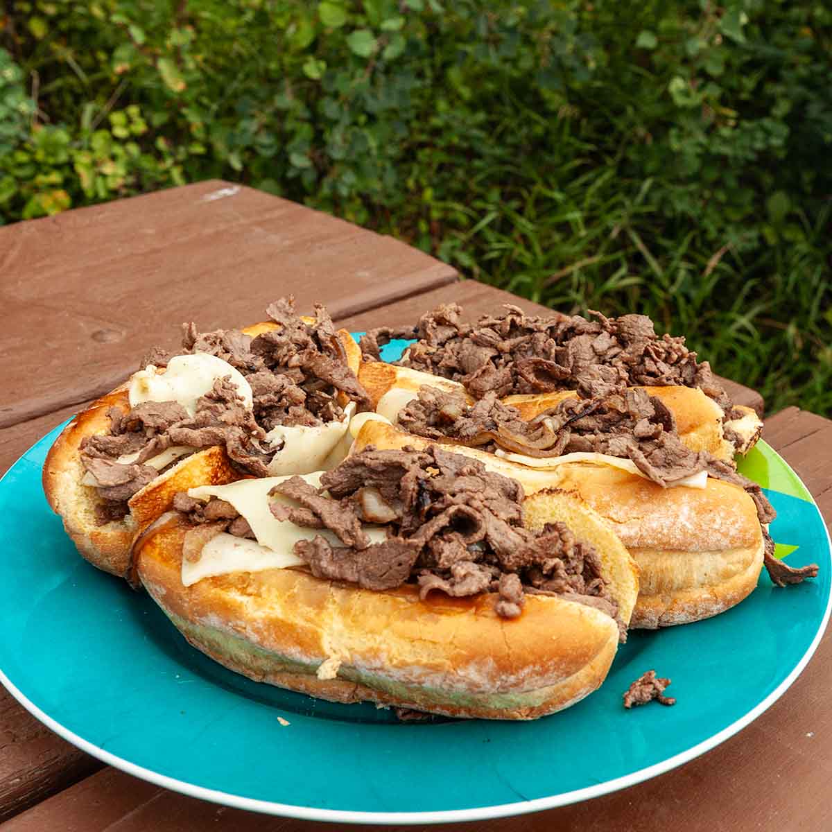 Four Blackstone cheesesteaks on a large plate on a picnic table.