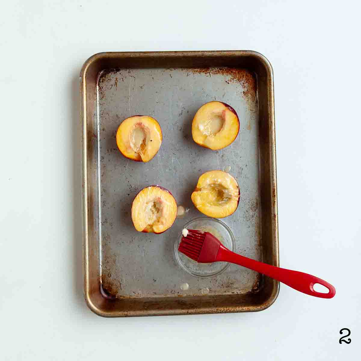 Four peach halves on a baking sheet with a dish of melted butter nearby.