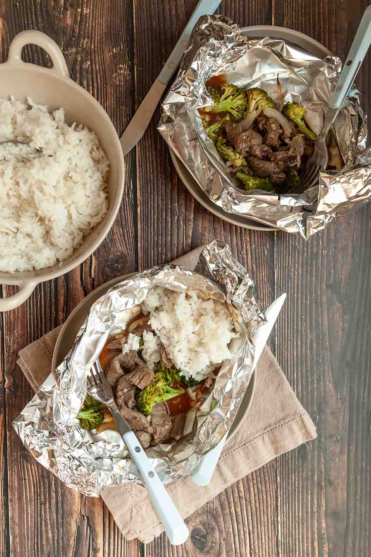 Two beef and broccoli foil packets with a bowl of rice on the side.