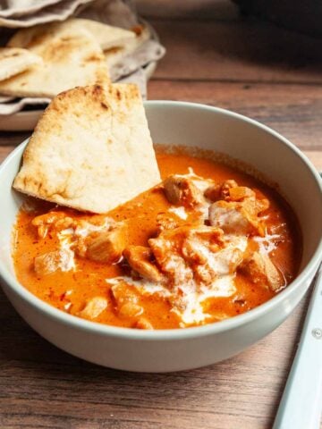 A bowl of butter chicken with a wedge of naan bread in it.