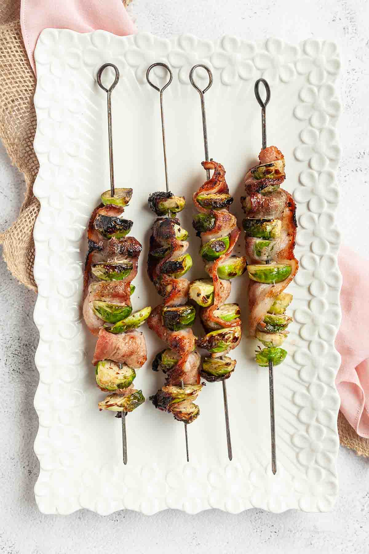 Four grilled Brussels sprouts and bacon skewers on a platter.