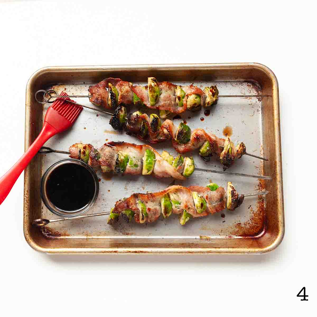 Four Brussels sprouts and bacon skewers on a sheet pan with a dish of balsamic vinegar beside them.