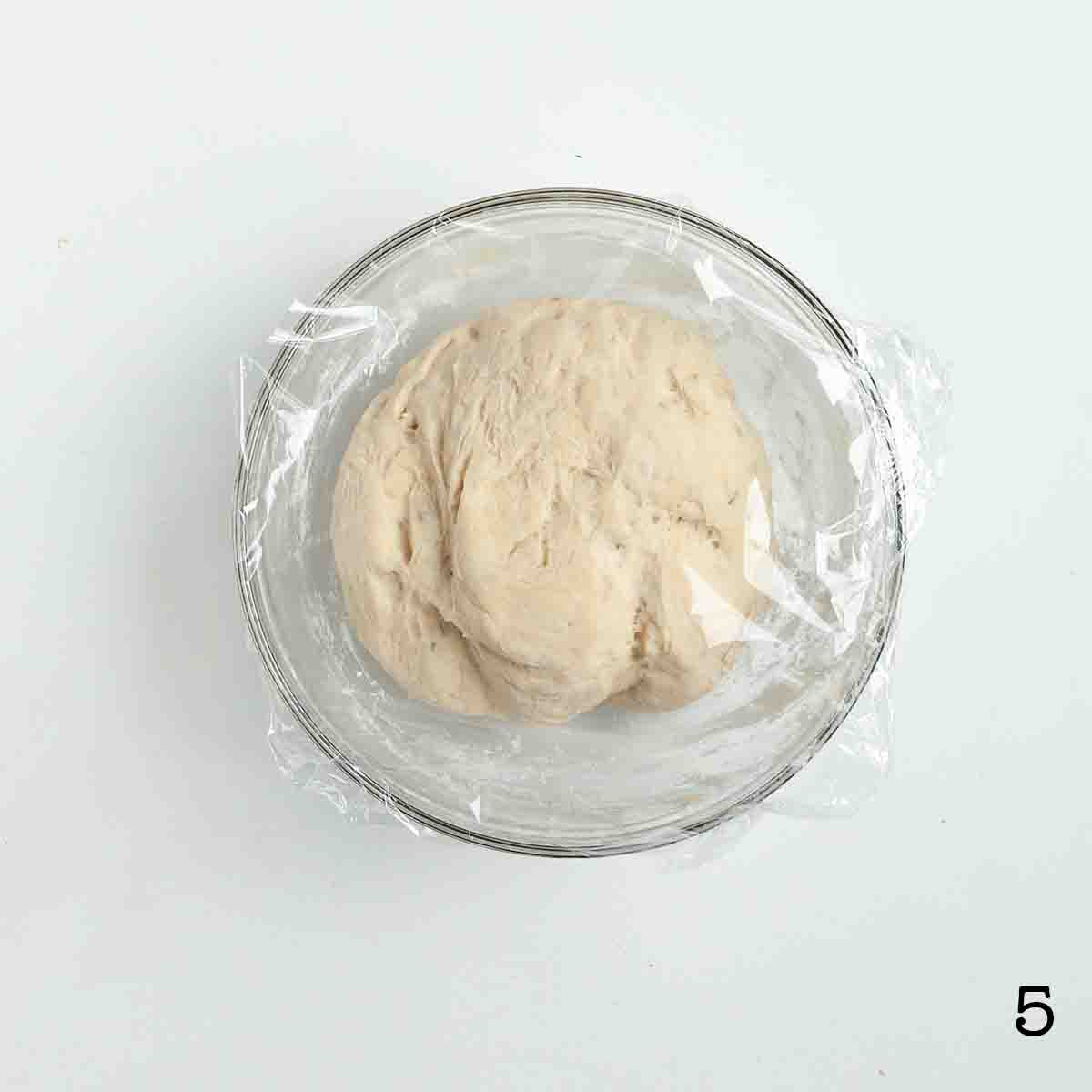 Risen bread dough covered in plastic wrap in a mixing bowl.