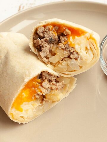 A breakfast burrito cut in half on a plate with hot sauce on the side.