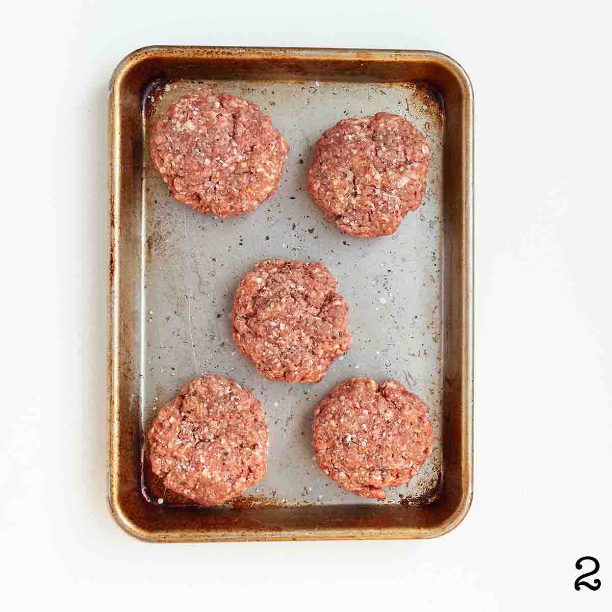Five uncooked burger patties on a baking sheet.