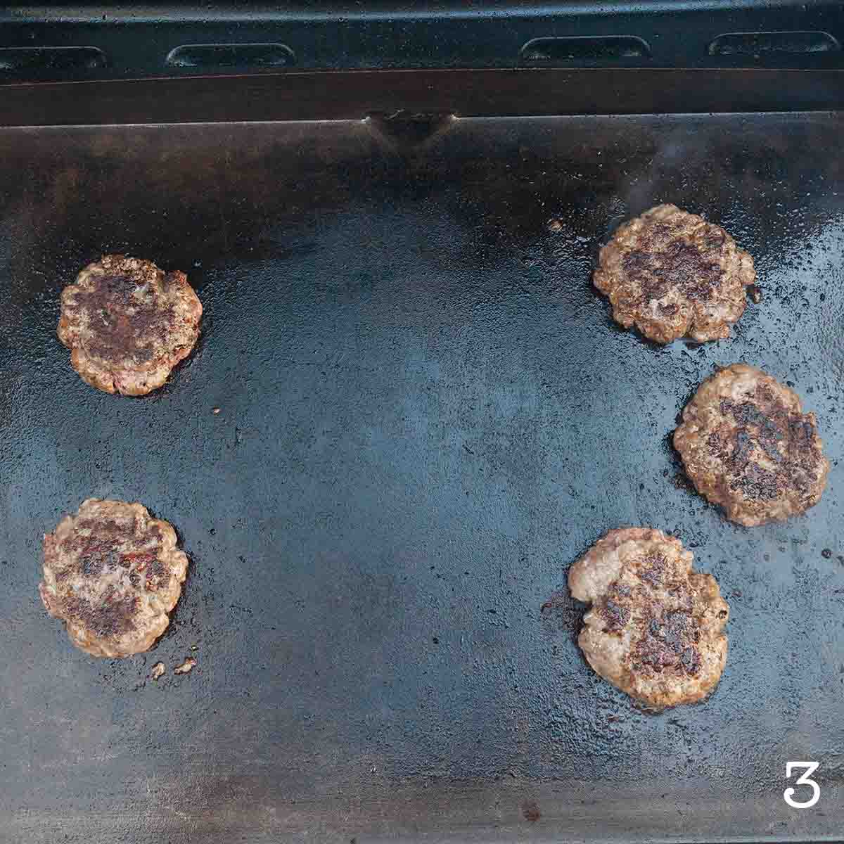Five burger patties cooking on an outdoor griddle.