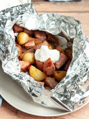 A foil packet filled with sausage and potato, topped with sour cream.