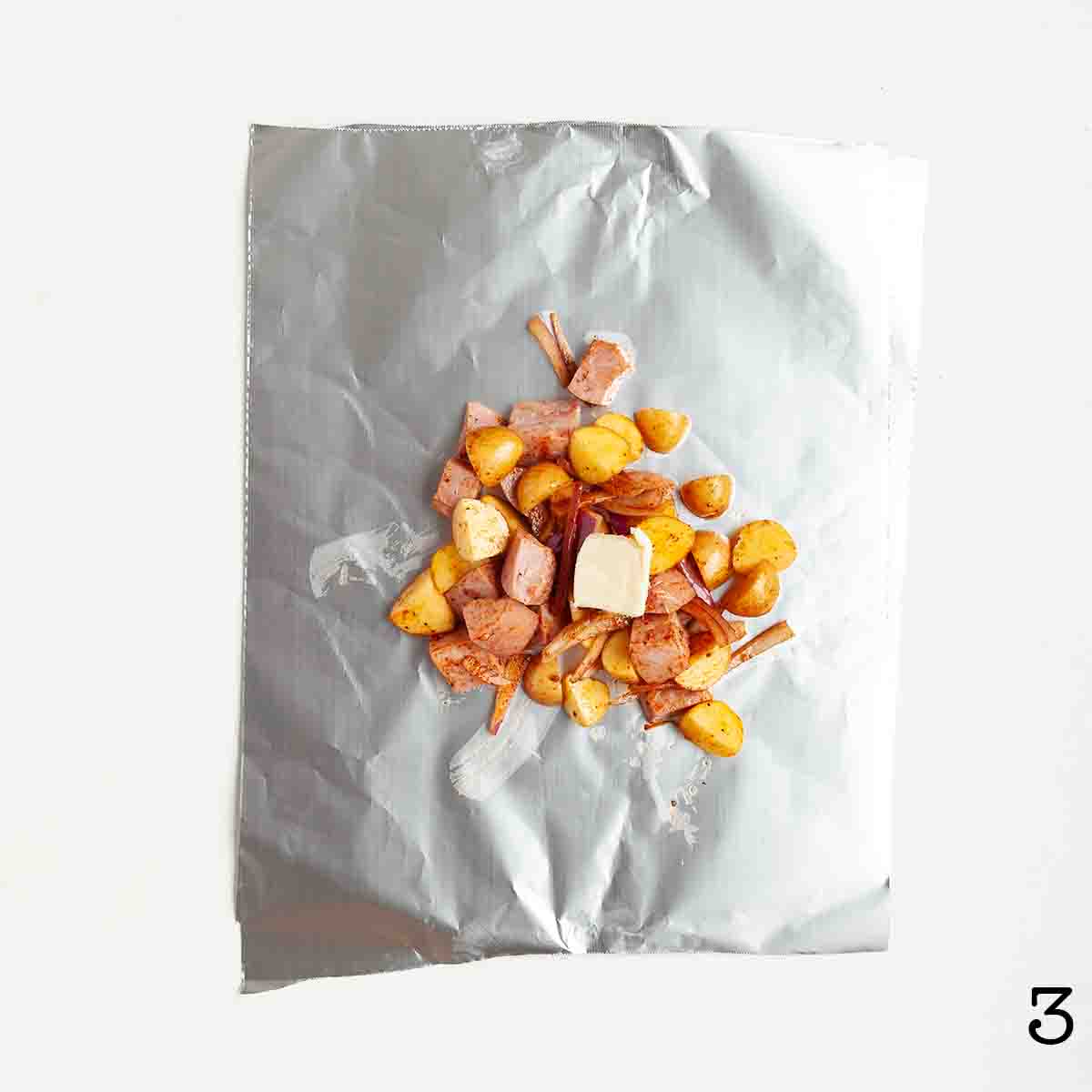 A portion of uncooked sausage, potatoes, and onion on foil, topped with a pat of butter.