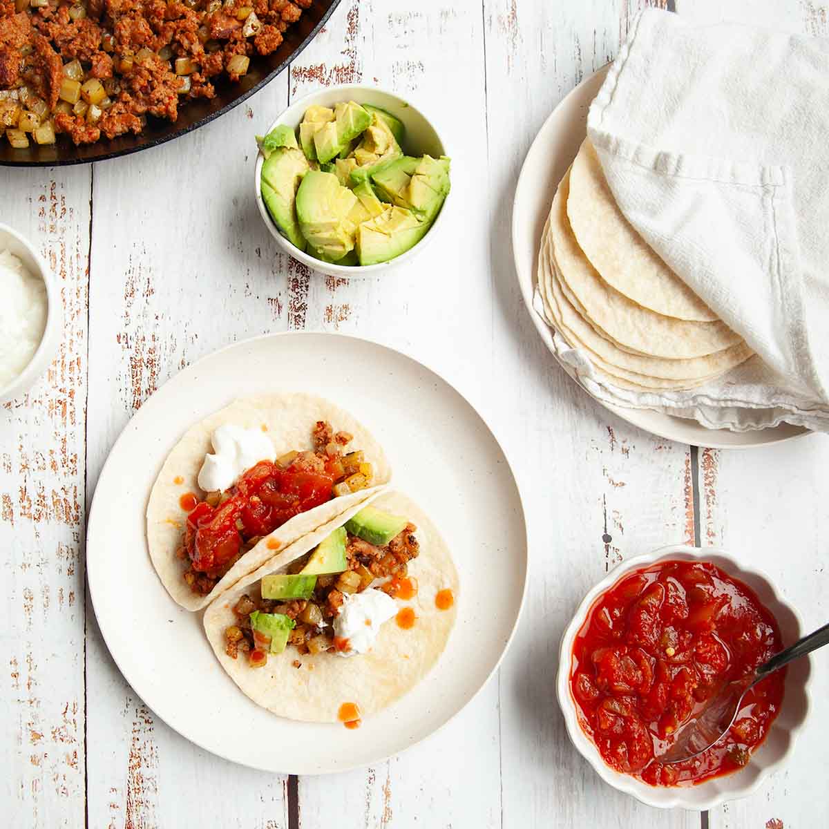 Two tacos with chorizo and potatoes, topped with salsa, sour cream, and avocado.