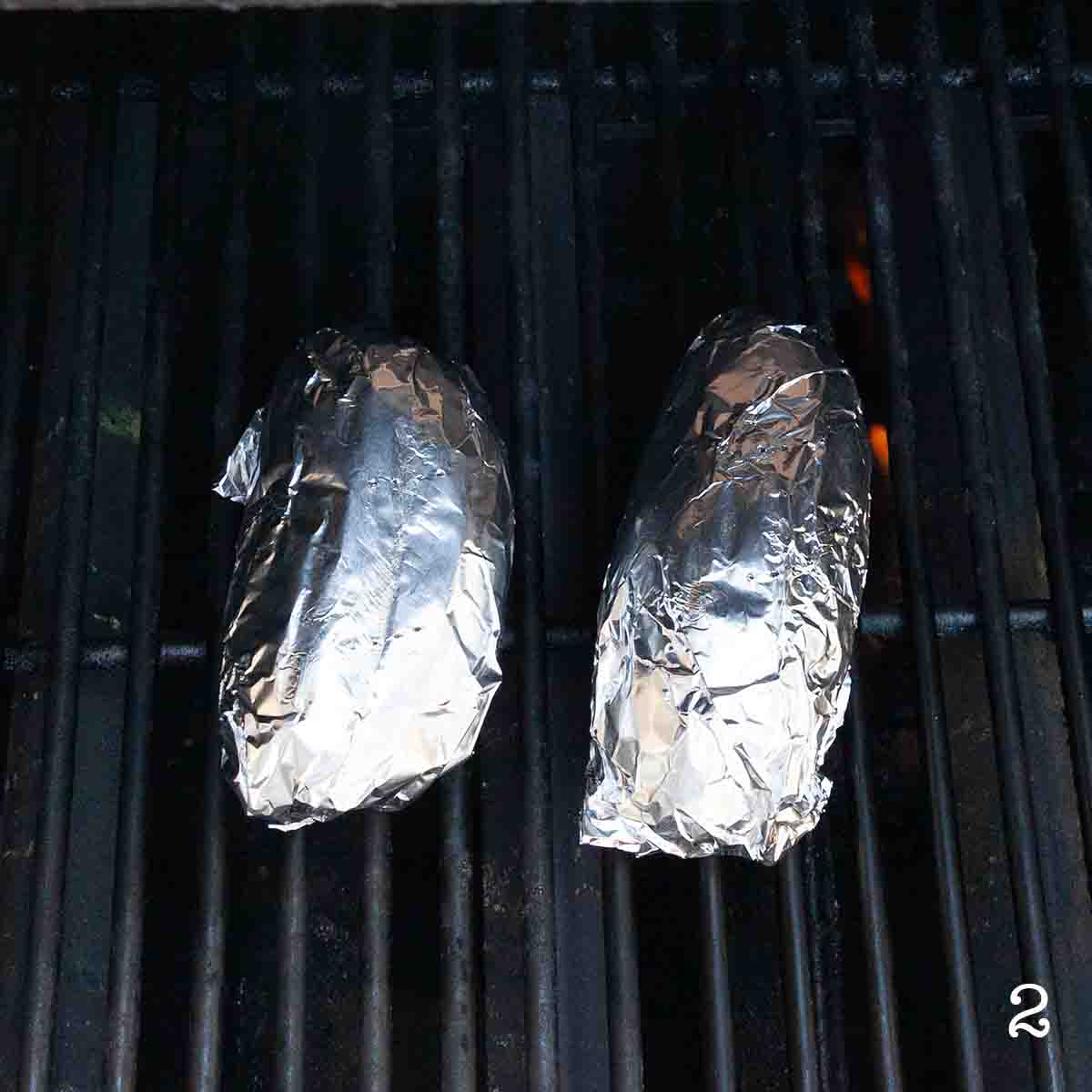 Two potatoes wrapped in foil cooking on the grill.