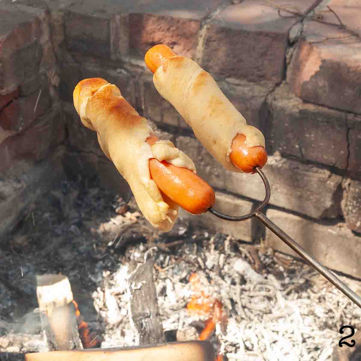 Two hot dogs wrapped in pastry over a campfire.