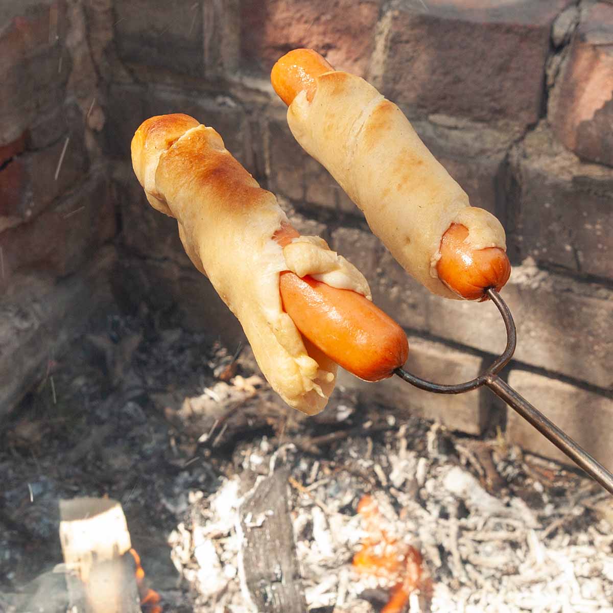Two hot dogs wrapped in pastry over a campfire.