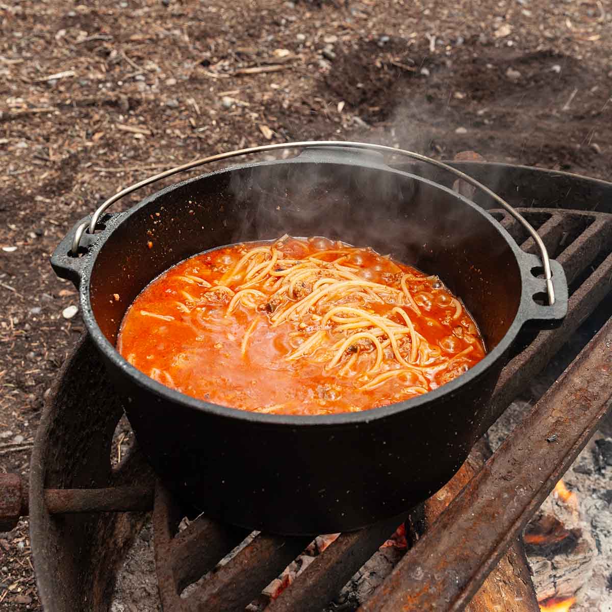 A Dutch oven filled with spaghetti and meat sauce over a campfire.