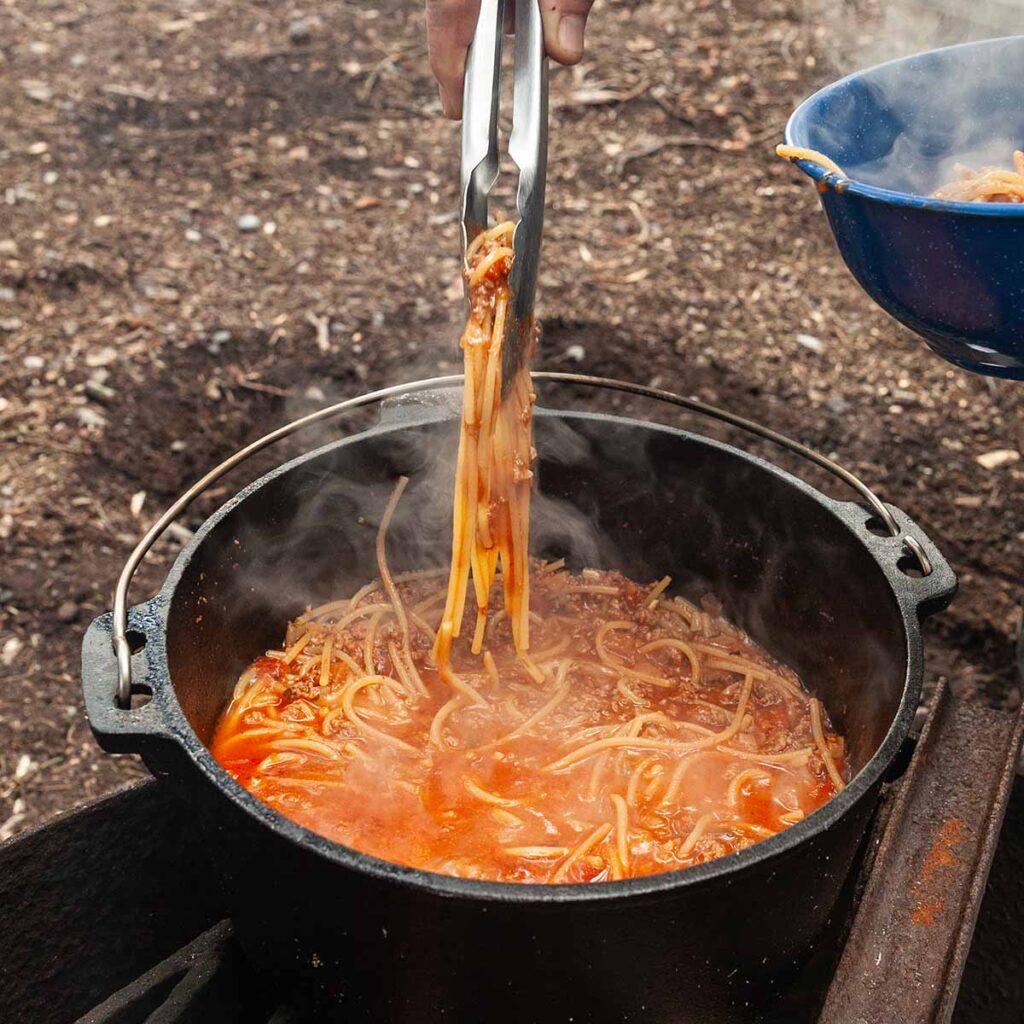 A person using tongs to pull spaghetti from a Dutch oven.