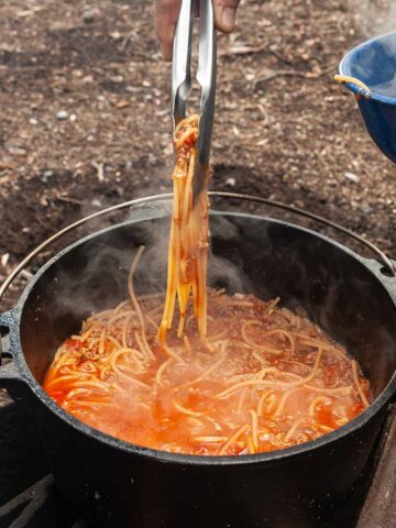 A person using tongs to pull spaghetti from a Dutch oven.