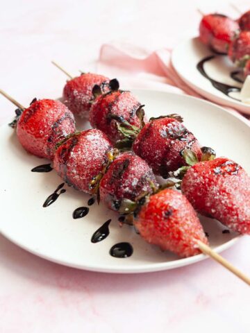 Skewers of grilled strawberries on a plate, drizzled with dark syrup.