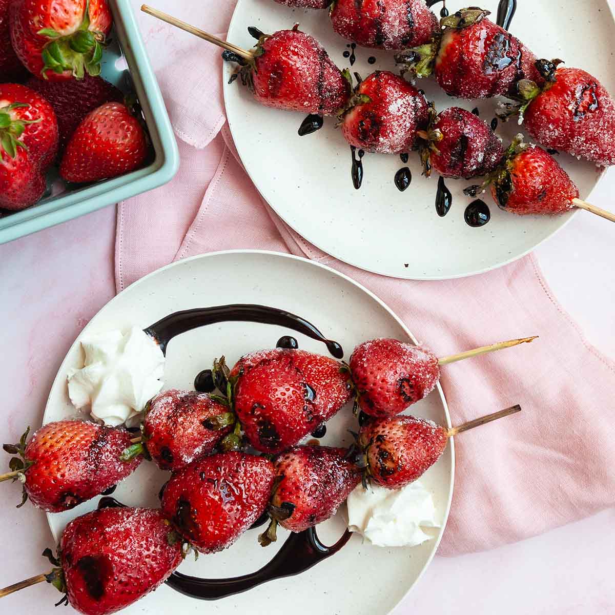 Skewers of grilled strawberries on a plate, drizzled with dark syrup.