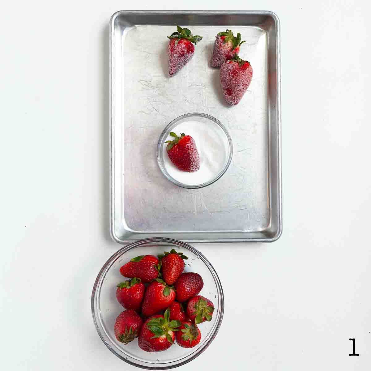 A bowl of strawberries and a bowl of sugar, with a strawberry being rolled in the sugar.