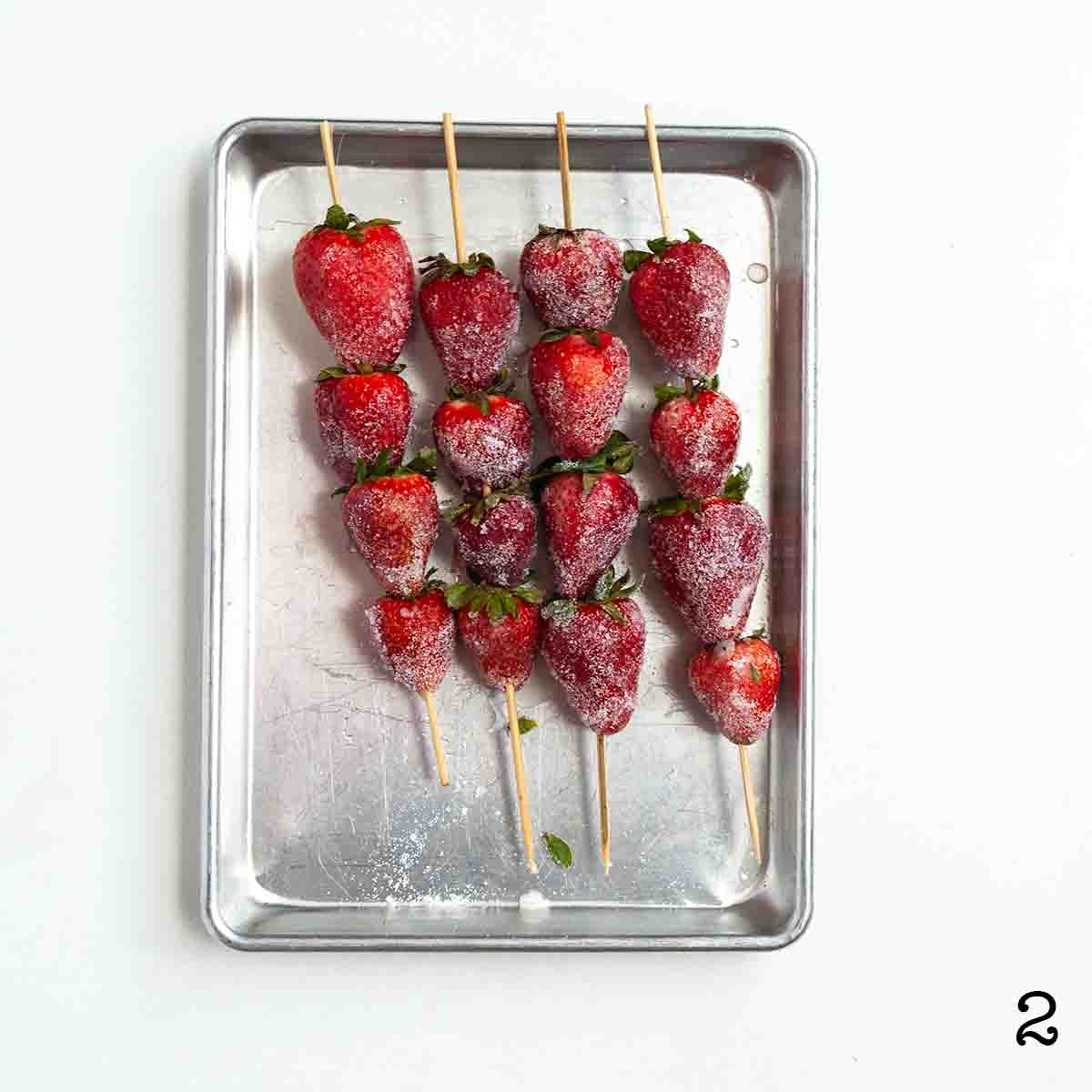 Four skewers of sugared strawberries on a baking sheet.