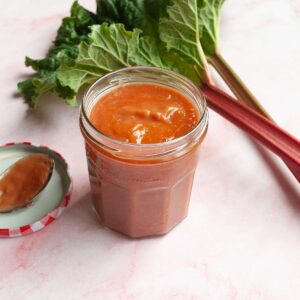 A jar of rhubarb bbq sauce with rhubarb stalks in the background.