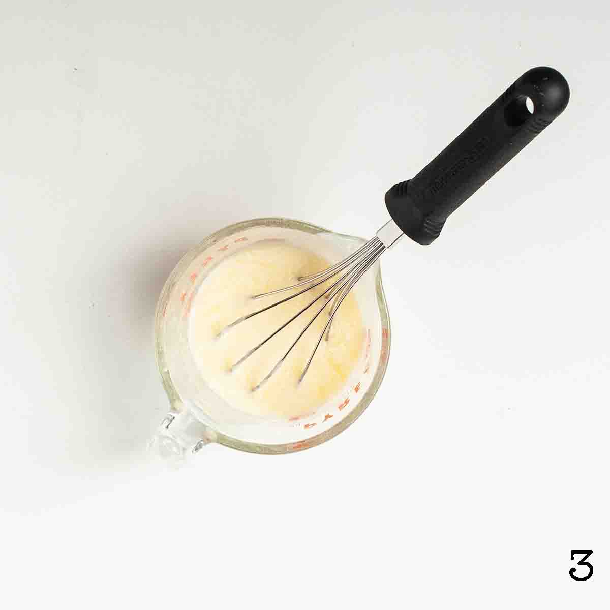 A measuring jug partially filled with liquid with a whisk resting inside.